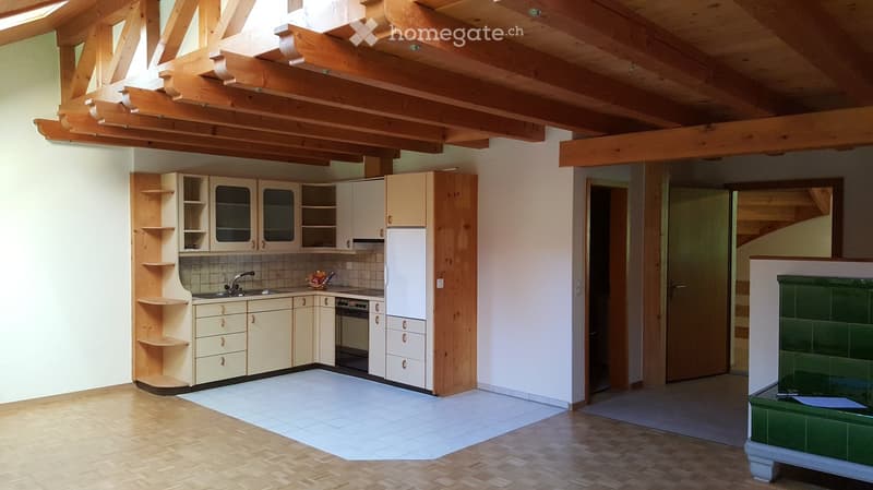 Bright kitchen with wooden beams on the ceiling, light-colored cabinets, a sink, a refrigerator, and an oven. The floor is wooden, and there is a green tiled fireplace on the right side.