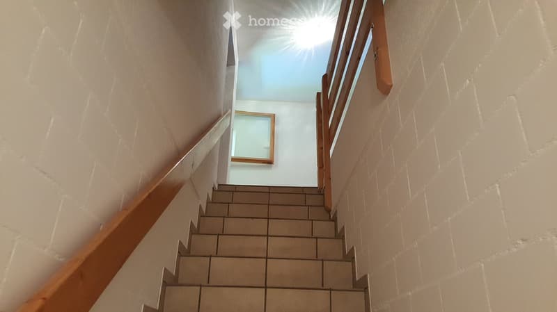 Staircase with beige tiled steps and wooden handrails, leading to a bright upper level with a skylight.