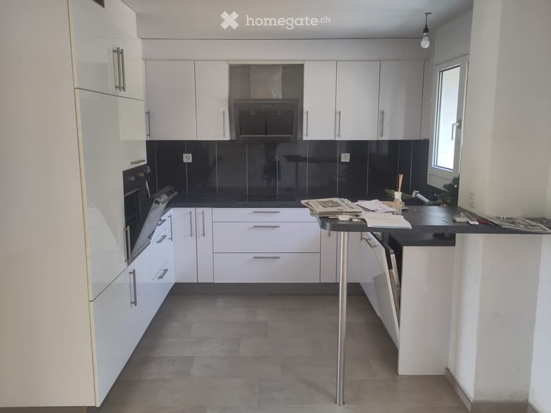 Modern kitchen with white cabinets, stainless steel appliances, and a black countertop. Features an island with a stool and various items on the counter.