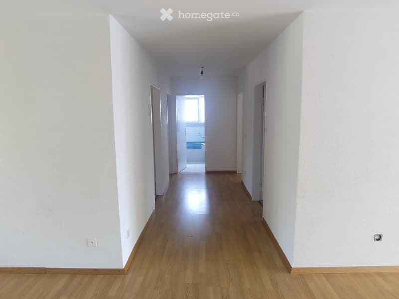 Empty hallway with light wooden flooring and white walls, leading to a partially visible bathroom at the end.