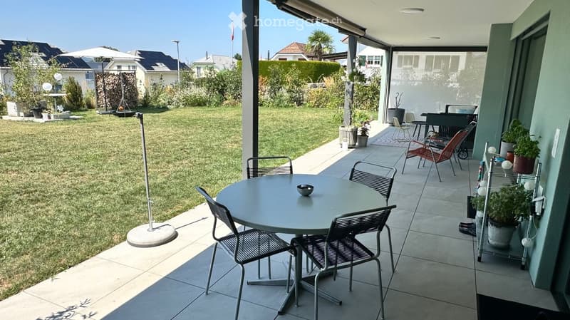 A well-maintained garden with a patio area featuring a round table and chairs, surrounded by greenery and potted plants. The patio is connected to a house with a covered walkway.