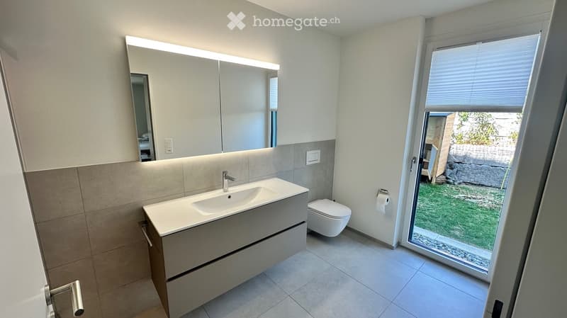 Modern bathroom with a white sink, toilet, large mirror with LED lighting, and a window with blinds overlooking a small garden.