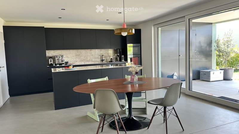 Modern kitchen with dark cabinets, a round wooden dining table, and six chairs. Features a pendant light, coffee machine, and an open view to the outdoor area.
