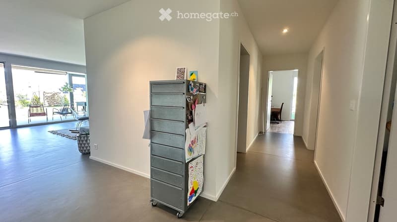 Bright hallway with white walls, wooden flooring, and a grey rolling cart with drawings attached. A large window at the end leads to a patio area with outdoor furniture.