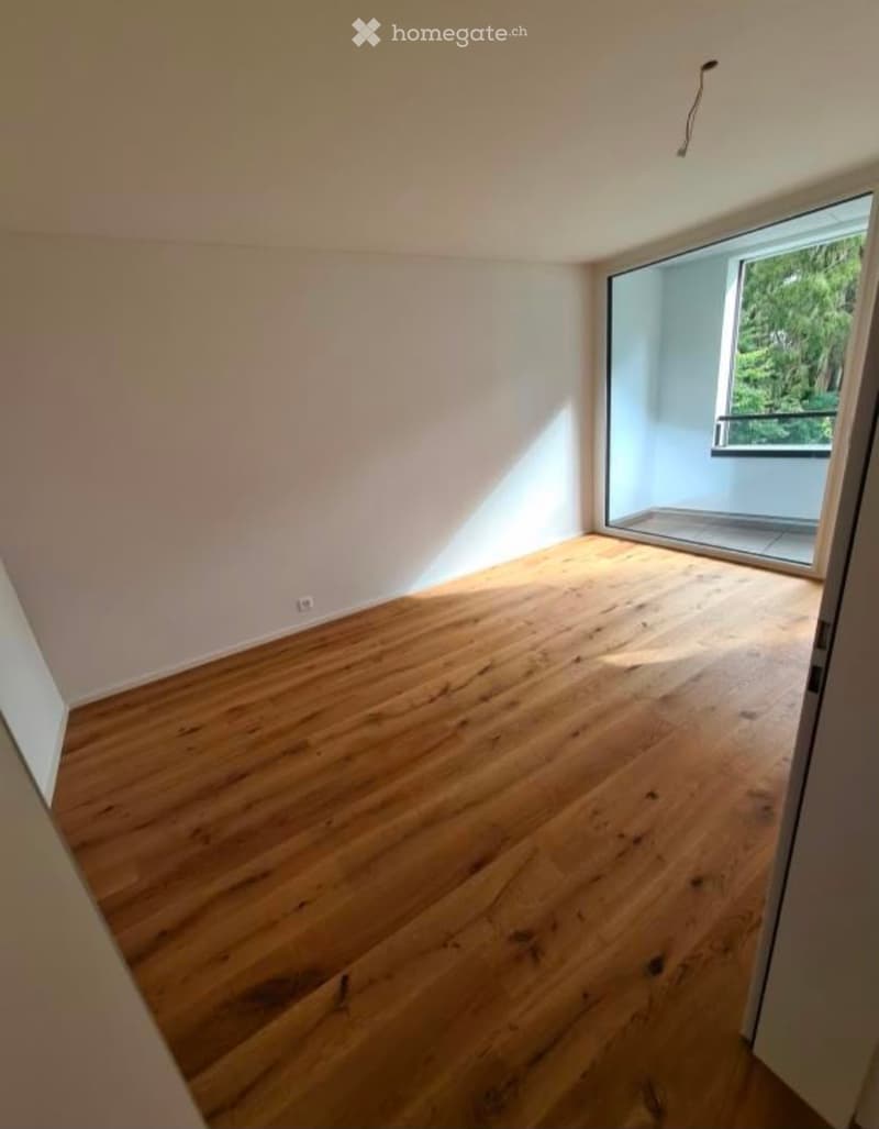 Empty room with wooden flooring and large window offering a view of greenery outside.