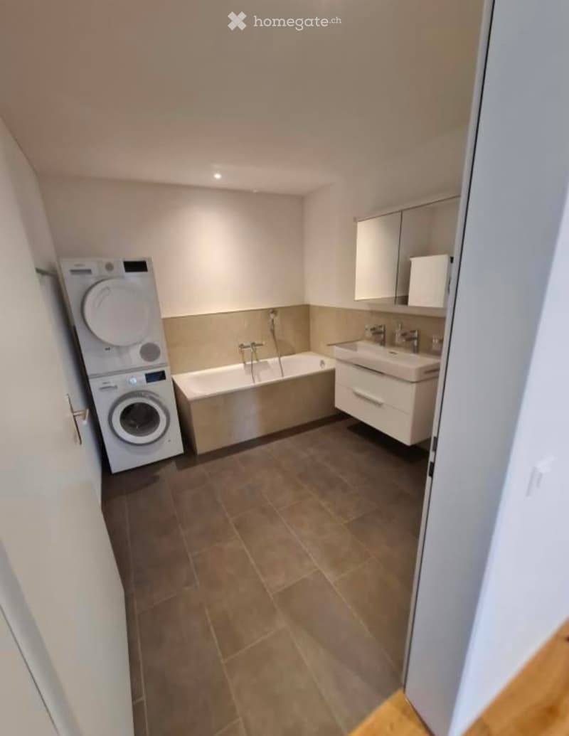 Modern bathroom with tiled floor, bathtub, sink, mirror, and a private washing machine.