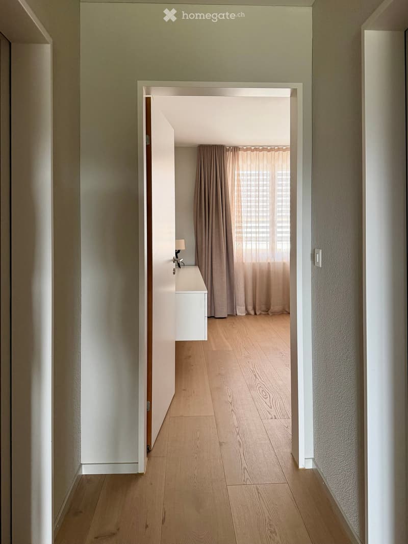 A modern hallway with light wooden flooring, white walls, and a small side table with a lamp. A doorway leads to a room with curtains and a window.