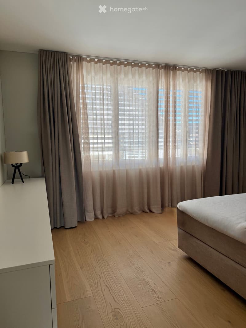 Modern bedroom with light wooden flooring, large window with curtains, a bed, and a bedside lamp.