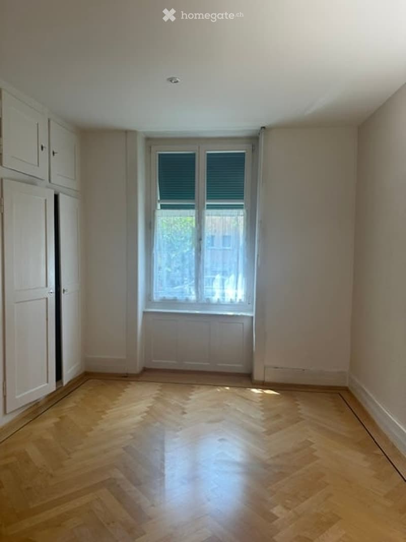Empty room with wooden herringbone flooring, white walls, and a large window with blinds. No furniture or equipment present.