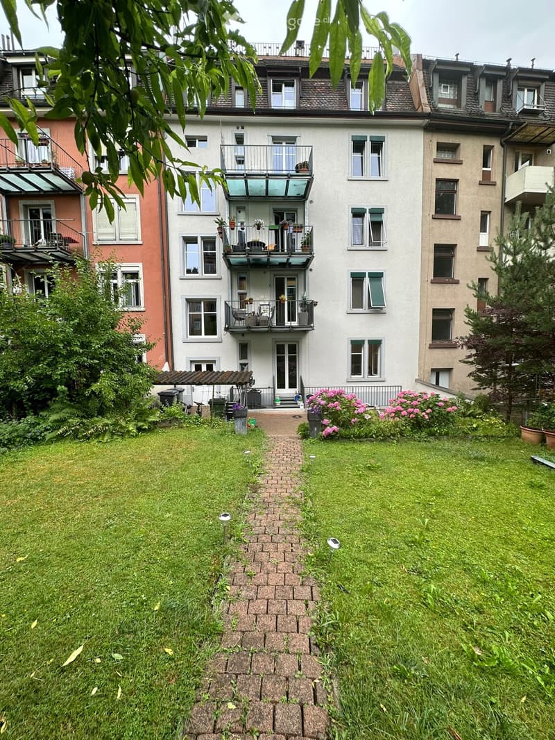Well-maintained garden with a brick pathway, green lawn, and various plants and flowers. The garden is bordered by a multi-story residential building with balconies and windows.