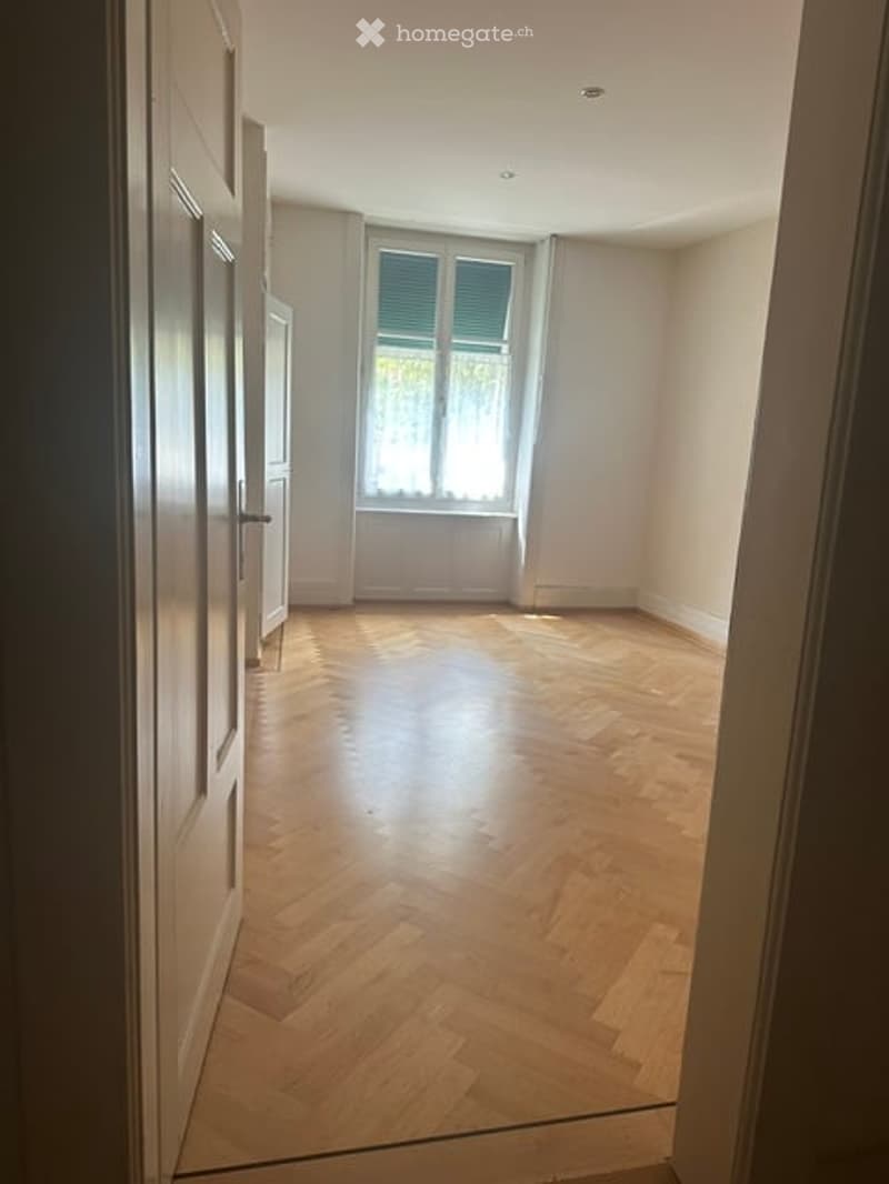 Empty room with wooden herringbone flooring, white walls, and a window with shutters. No furniture or equipment present.