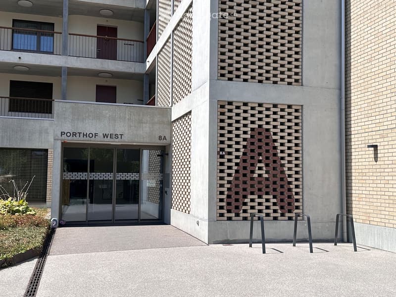 Modern building entrance labeled "PORTHOF WEST 8A" with glass doors, concrete walls, and a patterned brick facade.
