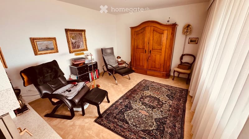 A well-furnished room with a leather sofa, a wooden cabinet, a patterned rug, and several chairs. The room features framed artwork on the walls and a small table with books and a lamp.