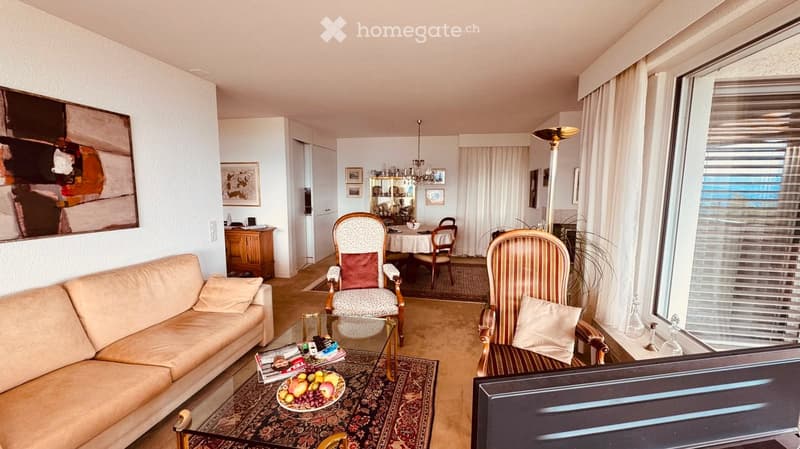 A well-furnished living room with a beige sofa, coffee table, two armchairs, a dining area with a table and chairs, a television, and decorative items including a fruit bowl and artwork.