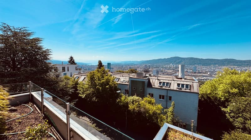 A rooftop terrace with a glass railing, overlooking a cityscape with buildings and trees under a clear blue sky.