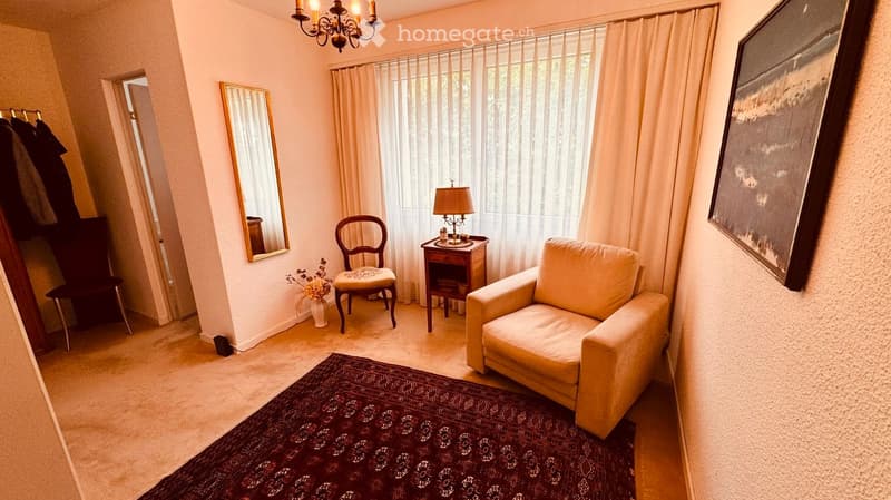 A cozy living room with a beige sofa, wooden chair, small wooden table with a lamp, a large mirror, and a patterned rug on the floor.