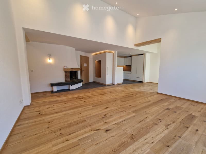 Open-plan living space with wooden flooring, white walls, fireplace, and adjacent kitchen area with white cabinets and appliances.