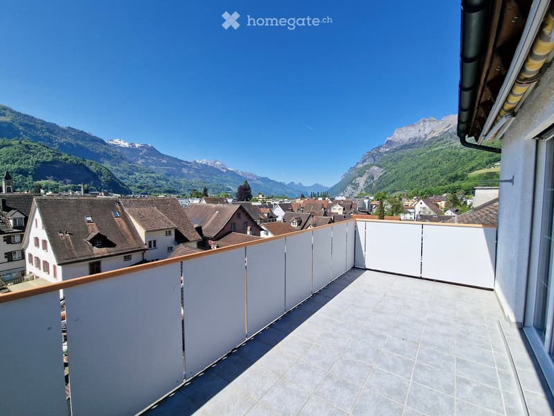 Balcony with tiled floor, white walls, wooden railing, overlooking a town with houses and distant mountains.