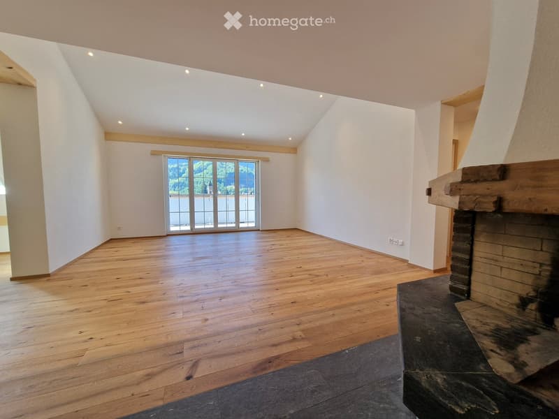 Empty room with wooden flooring, white walls, and a fireplace. Large window with a scenic view.