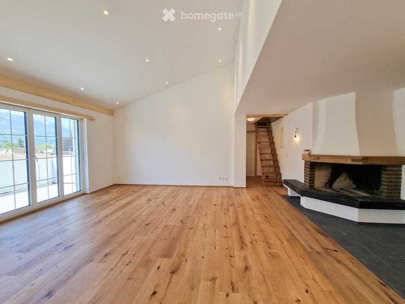 Empty living room with wooden flooring, white walls, large windows, and a fireplace with a wooden mantle.