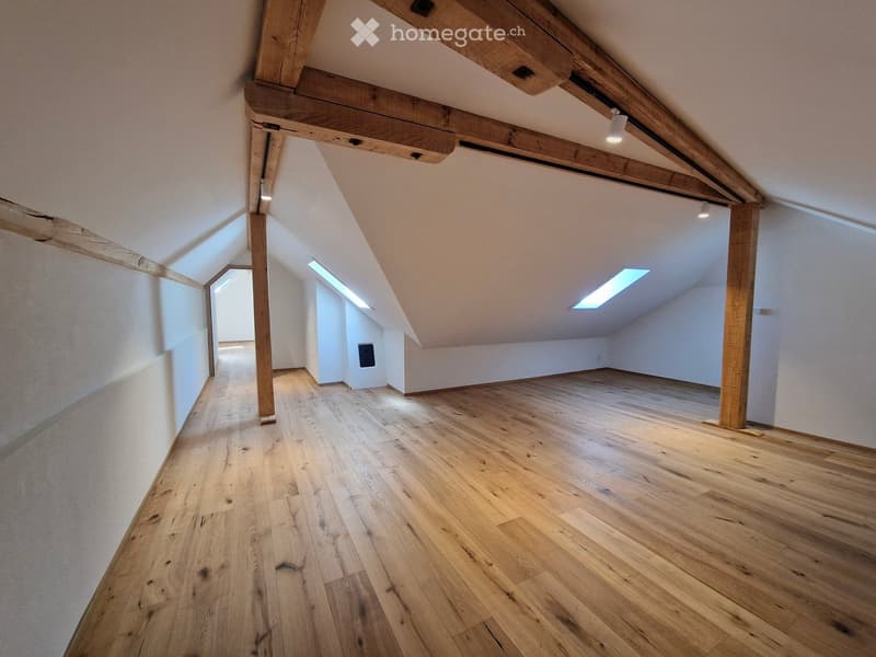 Empty attic space with wooden beams, wooden flooring, and skylight windows, featuring exposed wooden beams and white walls.