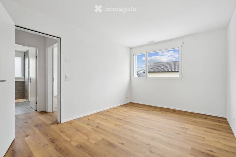 Empty room with white walls, wooden flooring, and a large window showing a view of a roof and sky. The room has a door leading to another space.