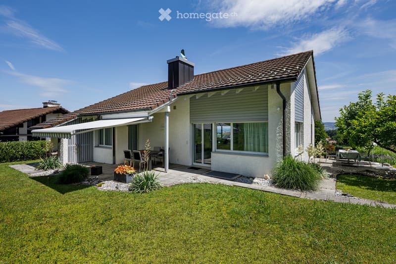 Single-story house with a tiled roof, white exterior, large windows, and a covered patio area. The front yard features a well-maintained lawn, garden beds, and outdoor seating.
