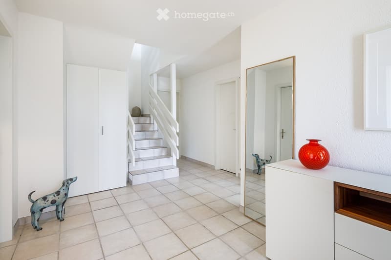Bright hallway with tiled floor, white walls, staircase leading upwards, and decorative items including a mirror, table with vase, and statues.