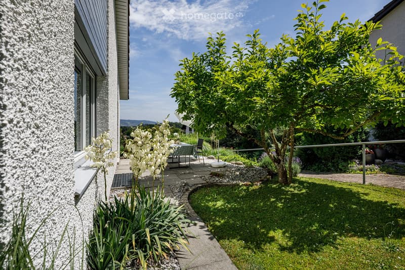 A well-maintained garden area with a patio, small table, chairs, and various plants and trees, adjacent to a modern house with a textured wall and large windows.