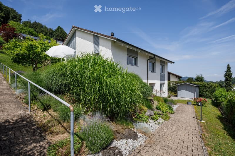 A two-story house with white walls and grey trim, surrounded by a well-maintained garden with various plants, shrubs, and a small shed. The house features multiple windows and a sloped roof.