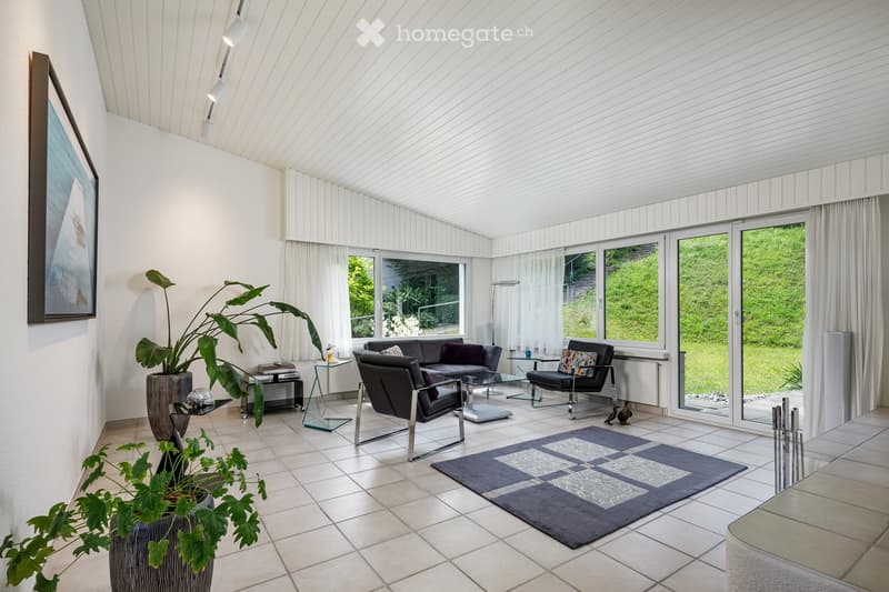 Modern living room with white walls, large windows, and a view of greenery outside. Features a dark grey sofa, black leather chairs, a glass coffee table, and a patterned rug.
