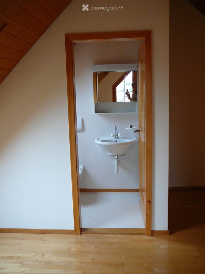Empty room with wooden flooring and white walls, featuring an open doorway leading to a small bathroom with a sink and mirror.