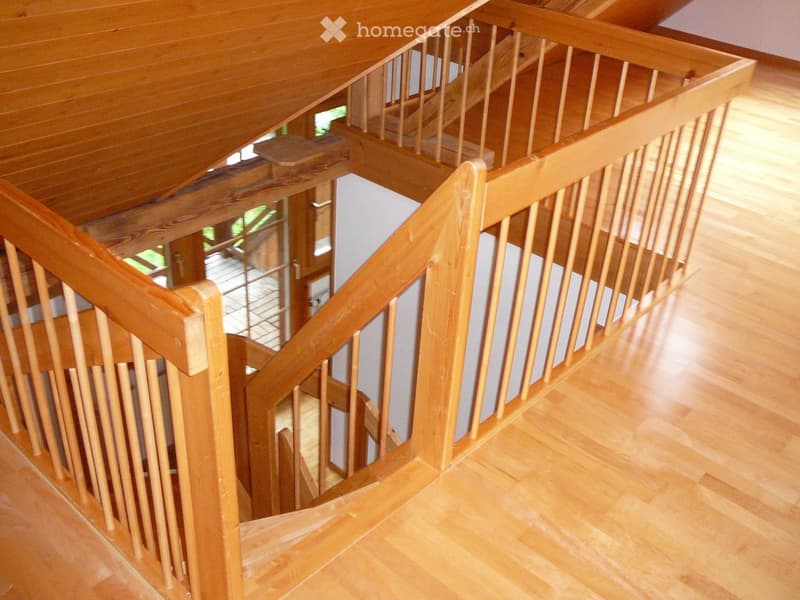 Wooden staircase with a polished wooden railing and balusters, leading to an upper level with visible wooden flooring and a slanted wooden ceiling.