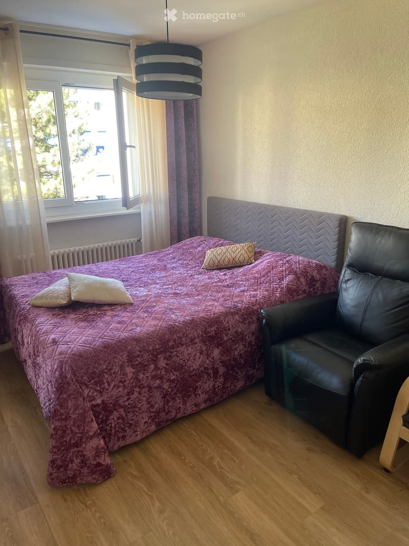 Cozy bedroom with a double bed covered in a pink quilt and decorative pillows, a black leather armchair, wooden flooring, and large windows with white curtains.