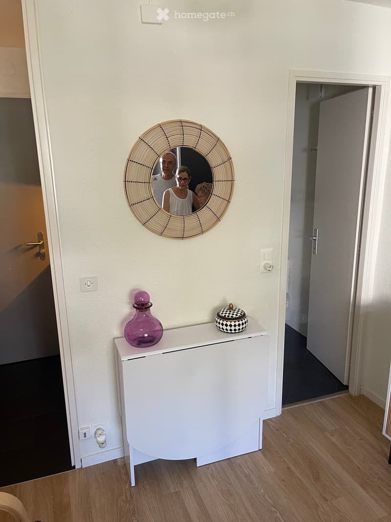 A hallway with a white cabinet topped with a purple vase and a decorative bowl, a round woven mirror on the wall, and a wooden floor.