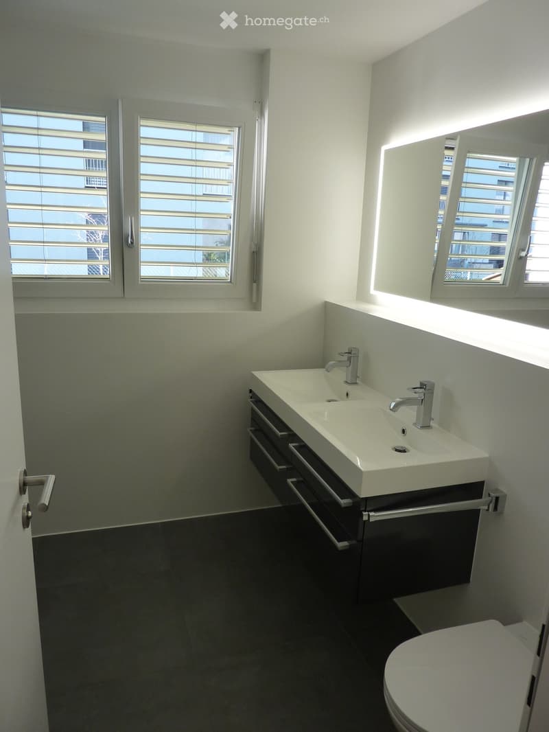 Modern bathroom with a white sink, chrome faucets, and a toilet. The room features large windows with blinds and a sleek, minimalist design.