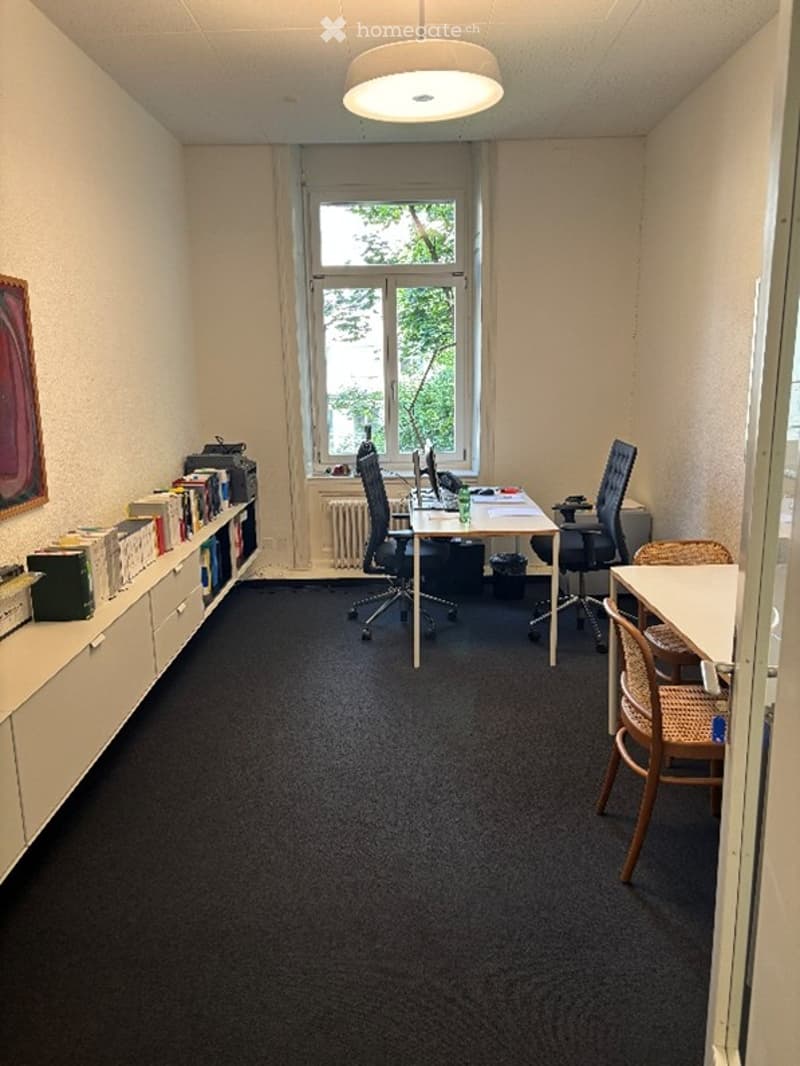 Office space with a desk, chairs, laptop, bookshelf, books, and a cup. The room has a large window with a view of greenery outside, a radiator below the window, and a picture frame on the wall.