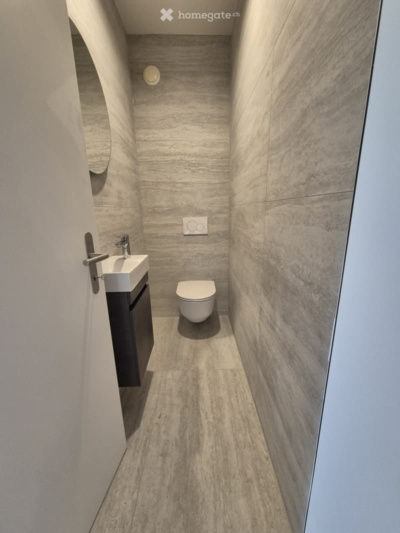 Modern, compact bathroom with marble tiles, a toilet, a sink with a faucet, and a dark cabinet.