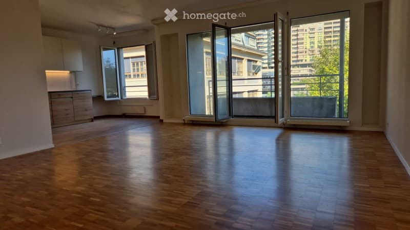 Empty room with wooden flooring, large windows, and a small kitchen area with cabinets.