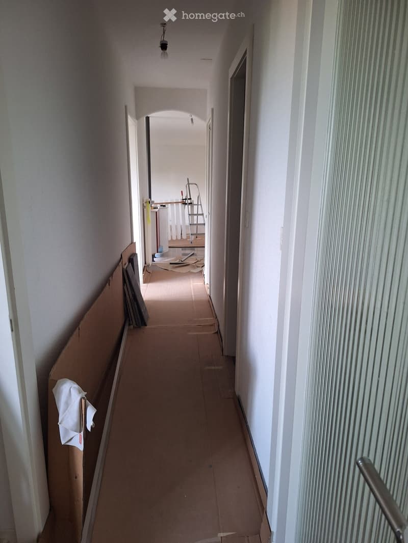 Empty hallway with white walls, wooden flooring, and a visible light fixture. There is a partially open door on the right leading to another room, and some construction materials are visible in the background.