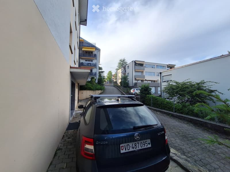 A narrow alleyway with a parked car, modern apartment buildings with balconies, greenery, and a clear sky.