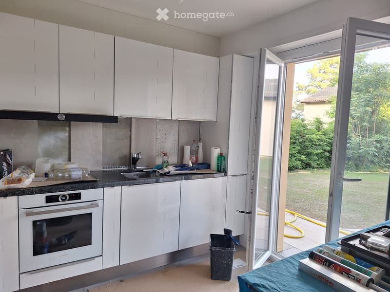 Modern kitchen with white cabinets, stainless steel oven, sink, and countertop. Open window reveals a green outdoor area.