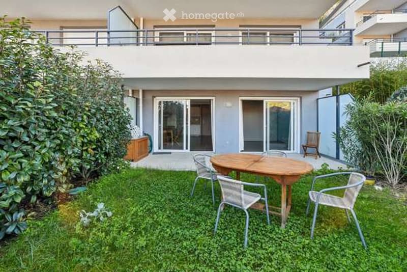 Garden area with a wooden table and chairs, surrounded by greenery and a modern building with a balcony above.