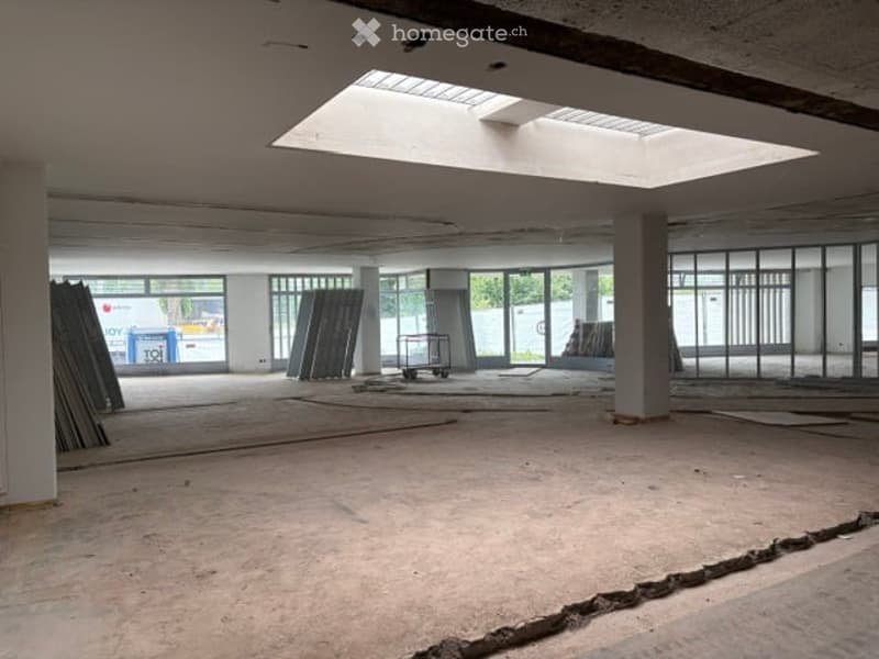 Empty, unfinished room with exposed concrete floor, large windows, and some construction materials visible.