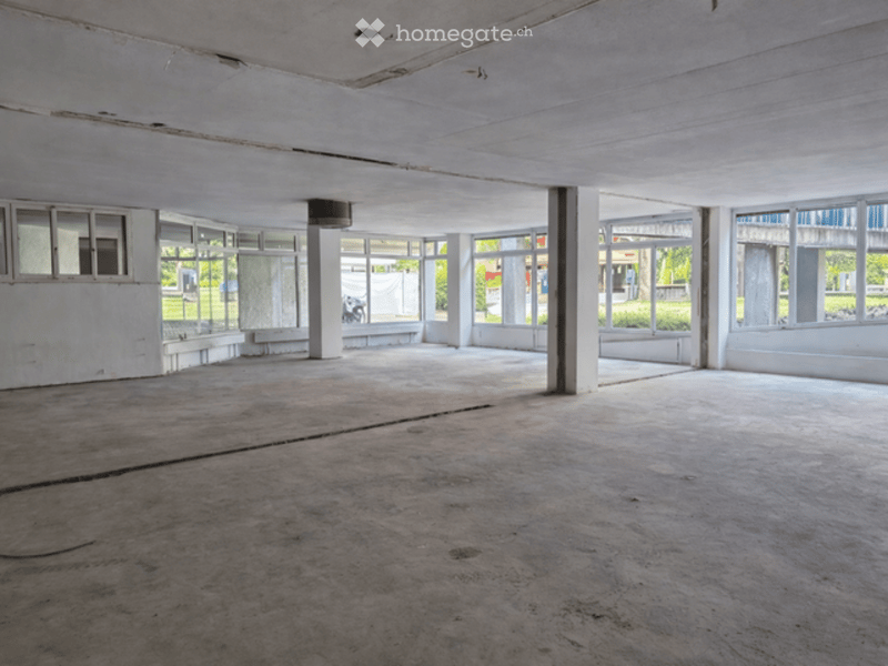 Empty room with concrete floor, white walls, large windows, and visible structural columns.