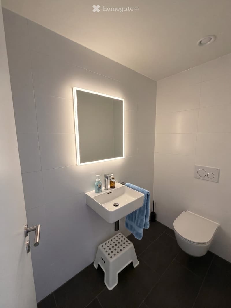 Modern bathroom with white tiles, a white sink, a mirror with LED lighting, a toilet, a small white stool, and a blue towel hanging on the side.