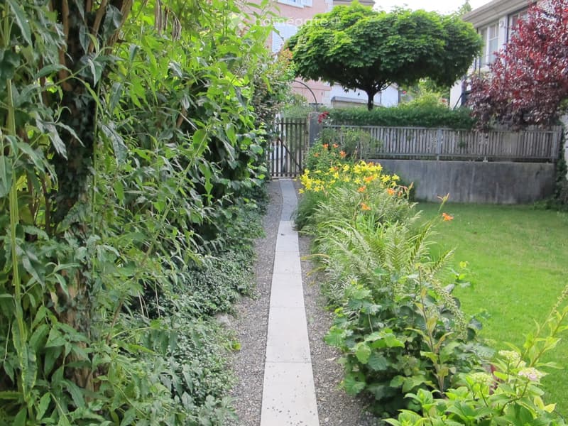 Well-maintained garden with a gravel pathway, various plants, shrubs, and a small lawn area. A wooden fence and gate are visible in the background.