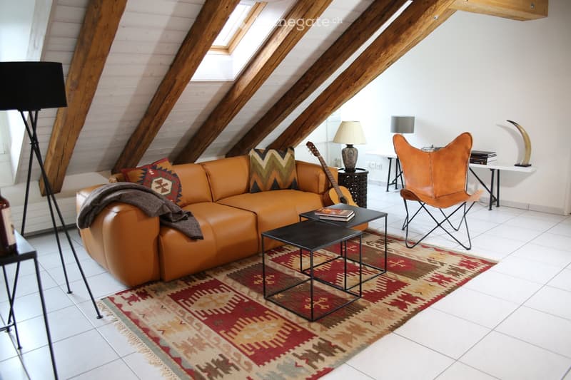 Living room with exposed wooden beams, white walls, and skylights. Features a brown leather sofa with patterned cushions, a guitar leaning against the sofa, a small black coffee table, a leather armchair, and a patterned rug.