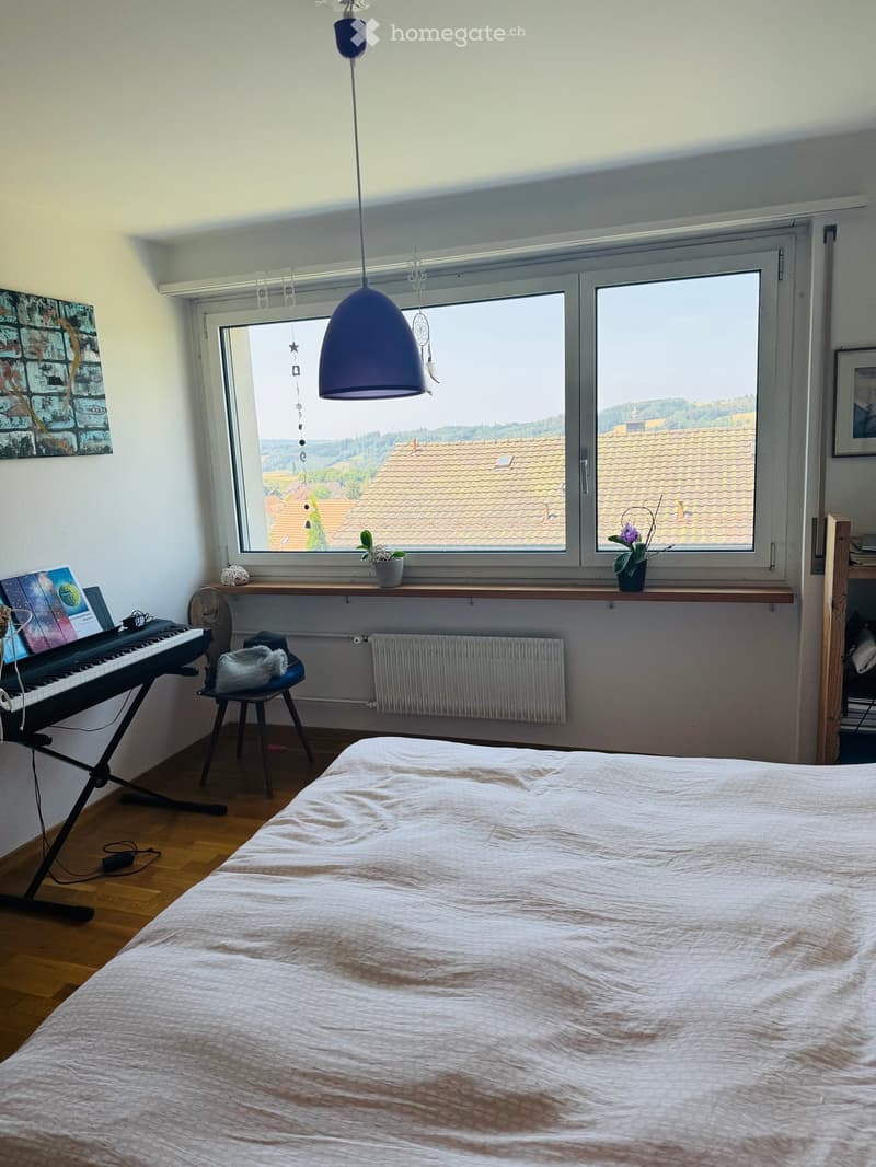 Bright bedroom with wooden flooring, large windows offering a view of rooftops, a bed with white bedding, a chair with a blanket, a small table with potted plants, a keyboard on a stand, and a hanging lamp.