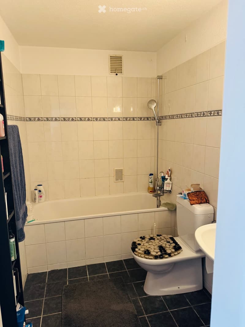Bathroom with a bathtub, toilet, sink, and shower. The walls are tiled with a decorative border, and there is a towel and several bottles on the shelf.
